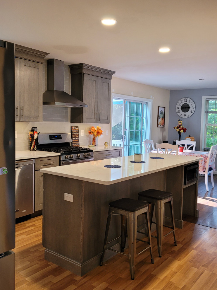 Example of a mid-sized transitional l-shaped light wood floor eat-in kitchen design in Boston with an undermount sink, shaker cabinets, gray cabinets, quartz countertops, white backsplash, subway tile backsplash, stainless steel appliances, an island and white countertops