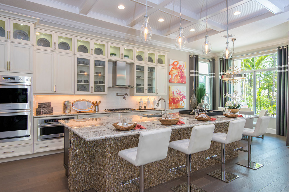 Photo of a tropical galley open plan kitchen in Other with white cabinets, white splashback, subway tile splashback, stainless steel appliances, medium hardwood floors, with island and glass-front cabinets.