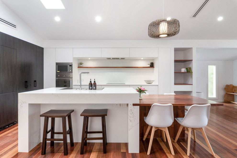 Inspiration for a contemporary l-shaped kitchen in Brisbane with flat-panel cabinets, white cabinets, white splashback, stainless steel appliances, dark hardwood floors, with island, brown floor, white benchtop and vaulted.