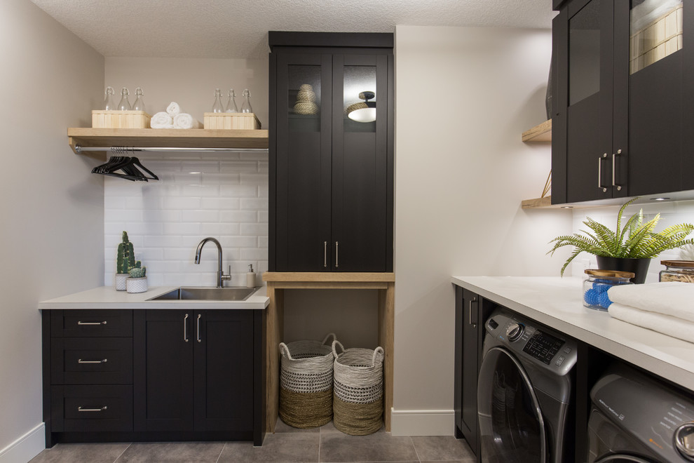 Inspiration for a large country l-shaped dedicated laundry room in Calgary with a drop-in sink, shaker cabinets, black cabinets, laminate benchtops, white walls, ceramic floors, an integrated washer and dryer, grey floor and grey benchtop.