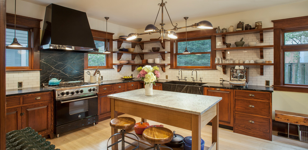 This is an example of an expansive arts and crafts l-shaped eat-in kitchen in Portland with raised-panel cabinets, soapstone benchtops, white splashback, subway tile splashback, black appliances, light hardwood floors, with island, an integrated sink and medium wood cabinets.