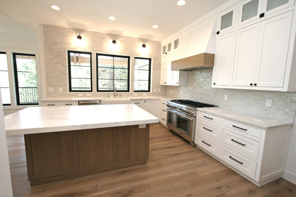 Photo of a large transitional u-shaped open plan kitchen in Seattle with a farmhouse sink, recessed-panel cabinets, white cabinets, marble benchtops, grey splashback, terra-cotta splashback, stainless steel appliances, medium hardwood floors, with island, beige floor and grey benchtop.