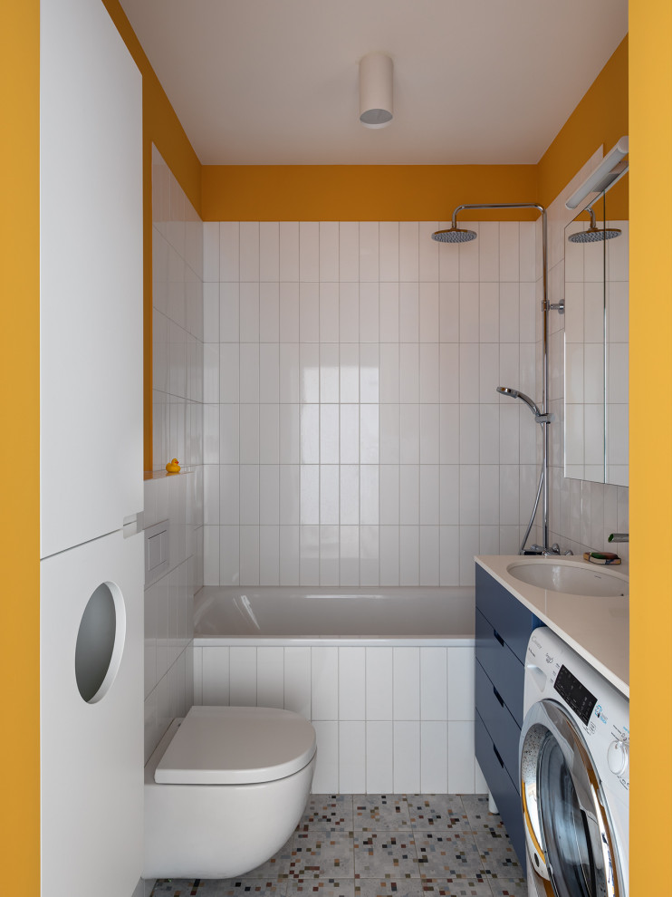 Exemple d'une petite salle de bain grise et jaune tendance avec un placard à porte plane, des portes de placard bleues, une baignoire en alcôve, un combiné douche/baignoire, WC suspendus, un carrelage blanc, un mur jaune, un lavabo encastré, un sol multicolore, aucune cabine, un plan de toilette blanc, buanderie, meuble simple vasque et meuble-lavabo suspendu.