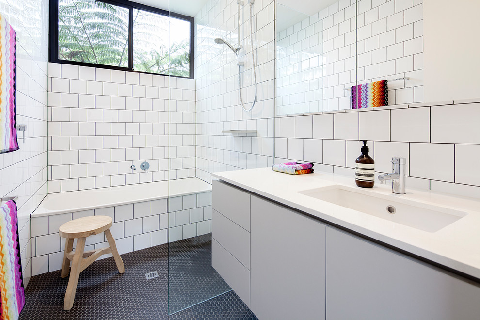 This is an example of a midcentury 3/4 bathroom in Melbourne with flat-panel cabinets, grey cabinets, a drop-in tub, a curbless shower, white tile, white walls, mosaic tile floors, an undermount sink, black floor, an open shower and white benchtops.