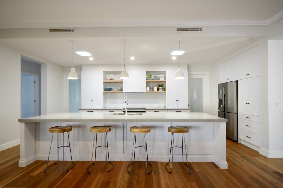 Photo of a large transitional l-shaped open plan kitchen in Sydney with an undermount sink, shaker cabinets, white cabinets, quartz benchtops, white splashback, subway tile splashback, stainless steel appliances, medium hardwood floors, with island, brown floor and grey benchtop.