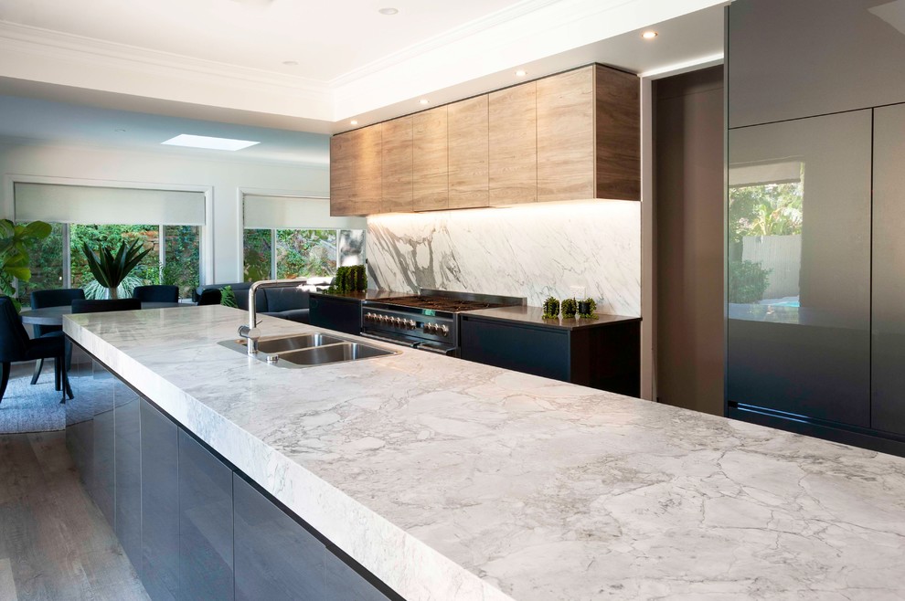 This is an example of a large contemporary galley open plan kitchen in Sydney with a drop-in sink, marble benchtops, stone slab splashback, black appliances and with island.