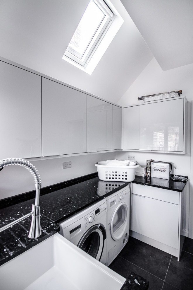 This is an example of a small contemporary u-shaped laundry cupboard in Surrey with a single-bowl sink, open cabinets, white cabinets, solid surface benchtops, white walls, porcelain floors, a side-by-side washer and dryer, black floor and black benchtop.