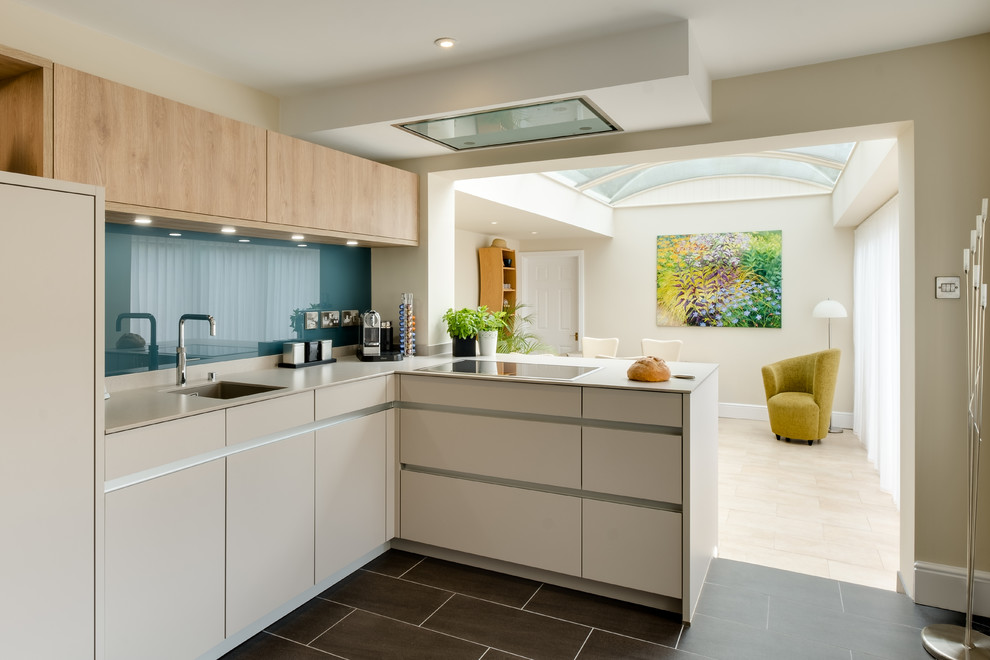 Photo of a mid-sized contemporary l-shaped open plan kitchen in Gloucestershire with flat-panel cabinets, quartzite benchtops, blue splashback, glass sheet splashback, panelled appliances, porcelain floors, an undermount sink, beige cabinets, a peninsula and black floor.