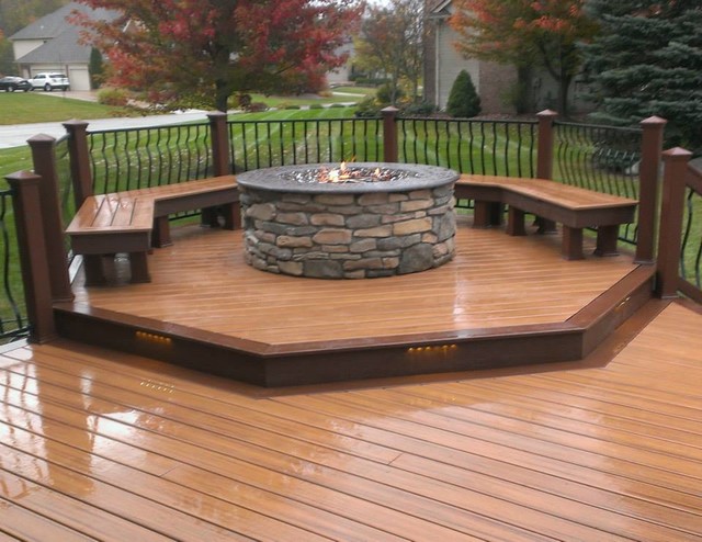 A stone fire pit on a wooden deck with built-in benches and integrated deck lighting, in a residential backyard.