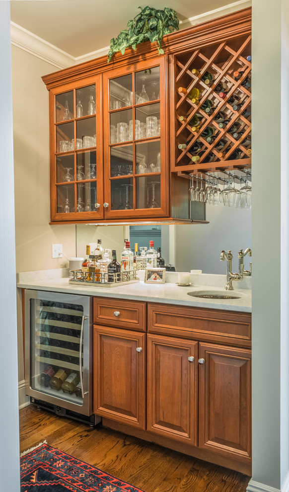 Photo of a small transitional single-wall wet bar in New York with an undermount sink, raised-panel cabinets, medium wood cabinets, solid surface benchtops, mirror splashback and medium hardwood floors.