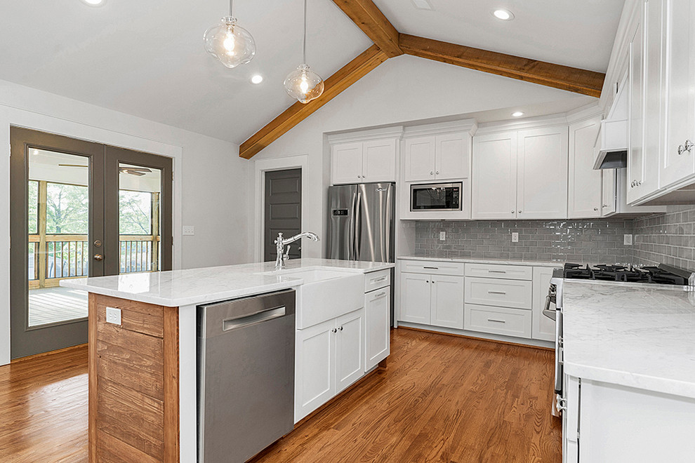 Mid-sized transitional l-shaped medium tone wood floor and brown floor open concept kitchen photo in Birmingham with a farmhouse sink, shaker cabinets, white cabinets, marble countertops, gray backsplash, subway tile backsplash, stainless steel appliances, an island and white countertops