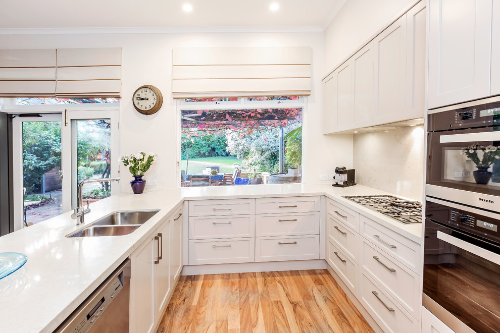This is an example of a transitional u-shaped eat-in kitchen in Adelaide with a double-bowl sink, shaker cabinets, white cabinets, grey splashback, medium hardwood floors, a peninsula, beige floor and white benchtop.