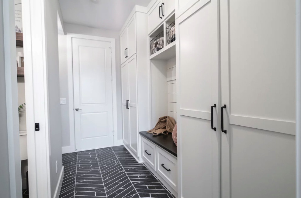 Design ideas for a mid-sized transitional galley utility room in Minneapolis with a farmhouse sink, shaker cabinets, white cabinets, quartz benchtops, white splashback, ceramic splashback, grey walls, ceramic floors, a stacked washer and dryer, black floor and black benchtop.