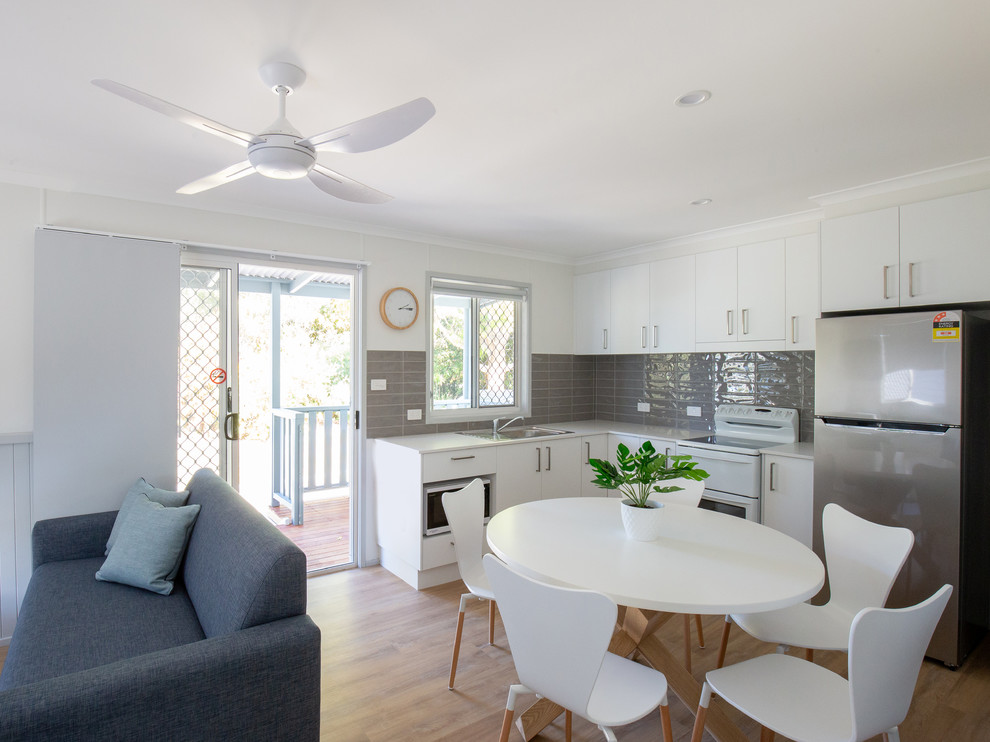 Photo of a small beach style l-shaped open plan kitchen in Gold Coast - Tweed with a single-bowl sink, white cabinets, laminate benchtops, grey splashback, ceramic splashback, stainless steel appliances, laminate floors, no island, brown floor and white benchtop.