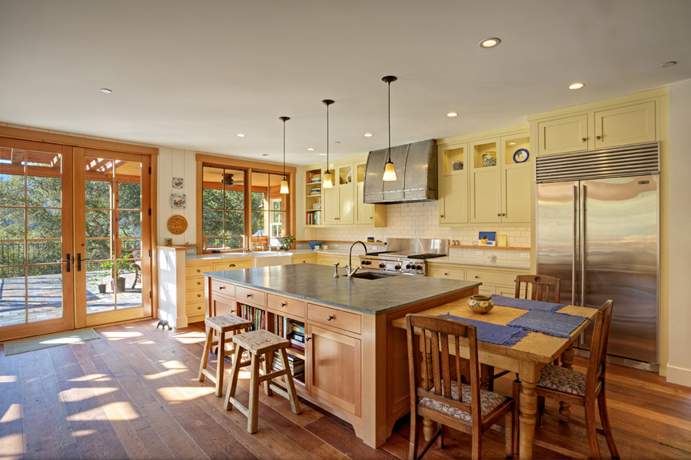 Design ideas for a mid-sized traditional l-shaped open plan kitchen in Austin with yellow cabinets, white splashback, subway tile splashback, stainless steel appliances, a farmhouse sink, shaker cabinets, soapstone benchtops, dark hardwood floors and with island.