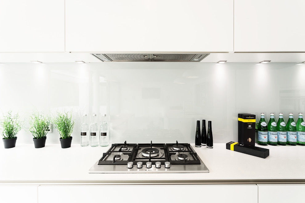 Photo of a large modern single-wall open plan kitchen in Hampshire with flat-panel cabinets, white cabinets, solid surface benchtops, grey splashback, glass sheet splashback, stainless steel appliances and with island.