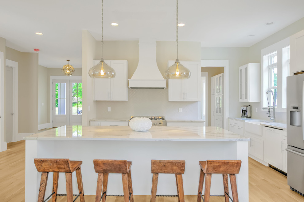 Inspiration for a mid-sized transitional l-shaped light wood floor open concept kitchen remodel with a farmhouse sink, shaker cabinets, white cabinets, stainless steel appliances, an island, quartzite countertops, white backsplash and ceramic backsplash