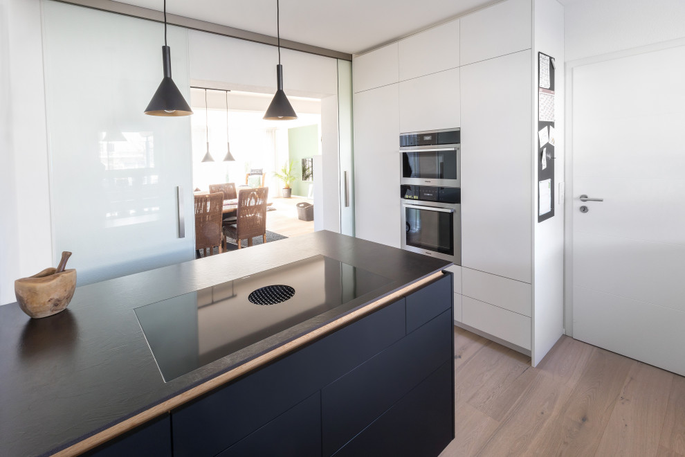 This is an example of a mid-sized contemporary galley separate kitchen in Stuttgart with white cabinets, white splashback, light hardwood floors, with island and black benchtop.