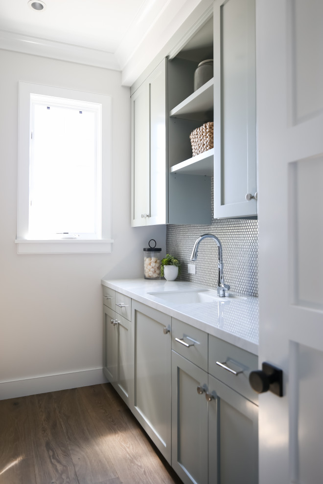 Mid-sized transitional galley dedicated laundry room in Orange County with an undermount sink, shaker cabinets, grey cabinets, white walls, medium hardwood floors, a side-by-side washer and dryer, beige floor and white benchtop.