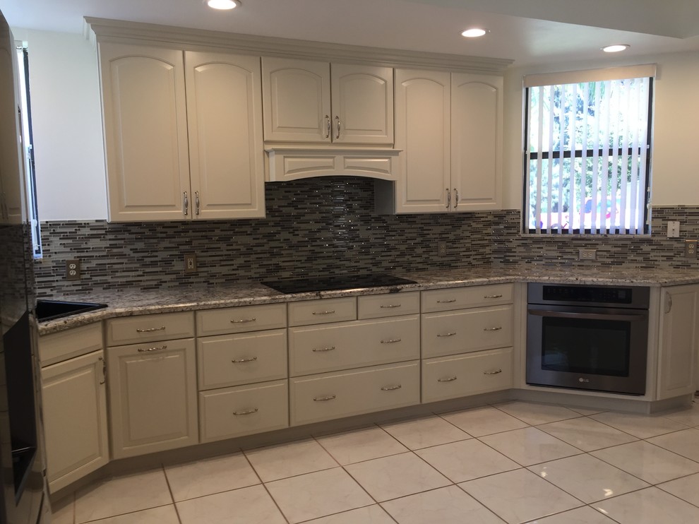 Mid-sized traditional l-shaped eat-in kitchen in Miami with a drop-in sink, raised-panel cabinets, beige cabinets, granite benchtops, glass sheet splashback, black appliances, porcelain floors, with island, multi-coloured splashback, beige floor and beige benchtop.