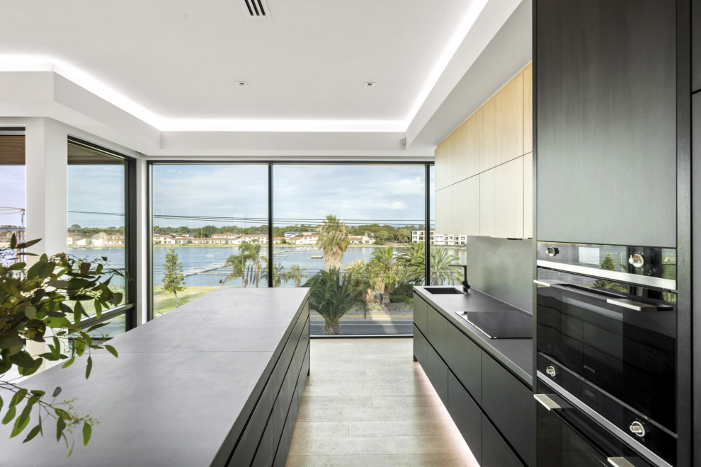 Large contemporary galley open plan kitchen in Adelaide with an integrated sink, flat-panel cabinets, black cabinets, grey splashback, black appliances, light hardwood floors, with island, beige floor and grey benchtop.