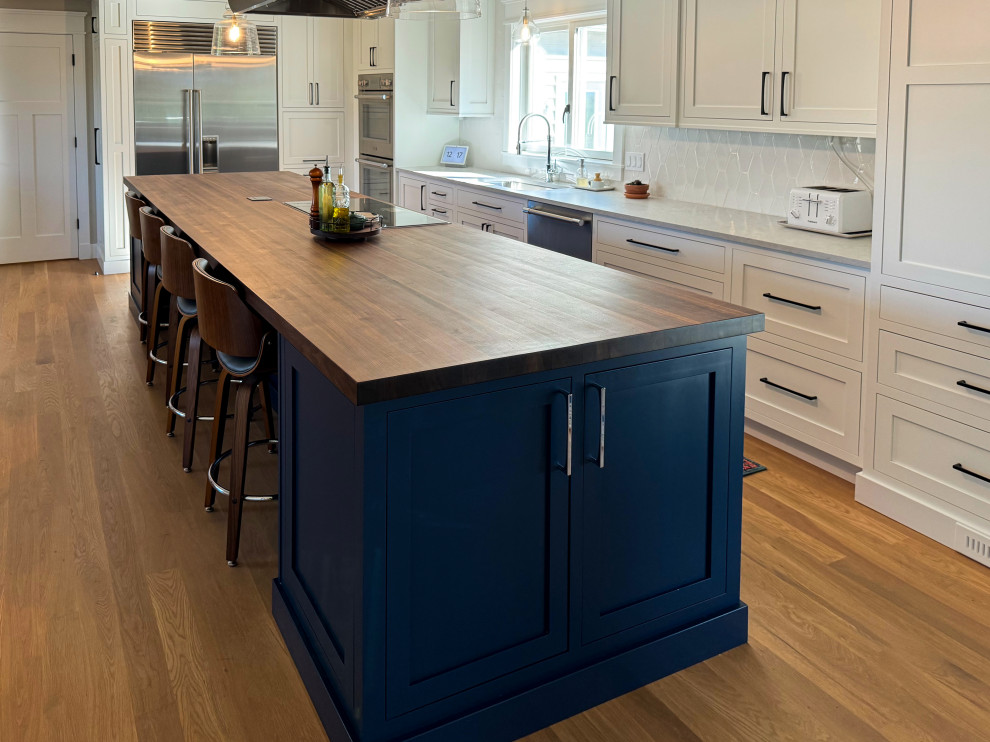 Mid-sized transitional l-shaped medium tone wood floor and brown floor eat-in kitchen photo with an undermount sink, shaker cabinets, wood countertops, white backsplash, an island, brown countertops, blue cabinets and stainless steel appliances