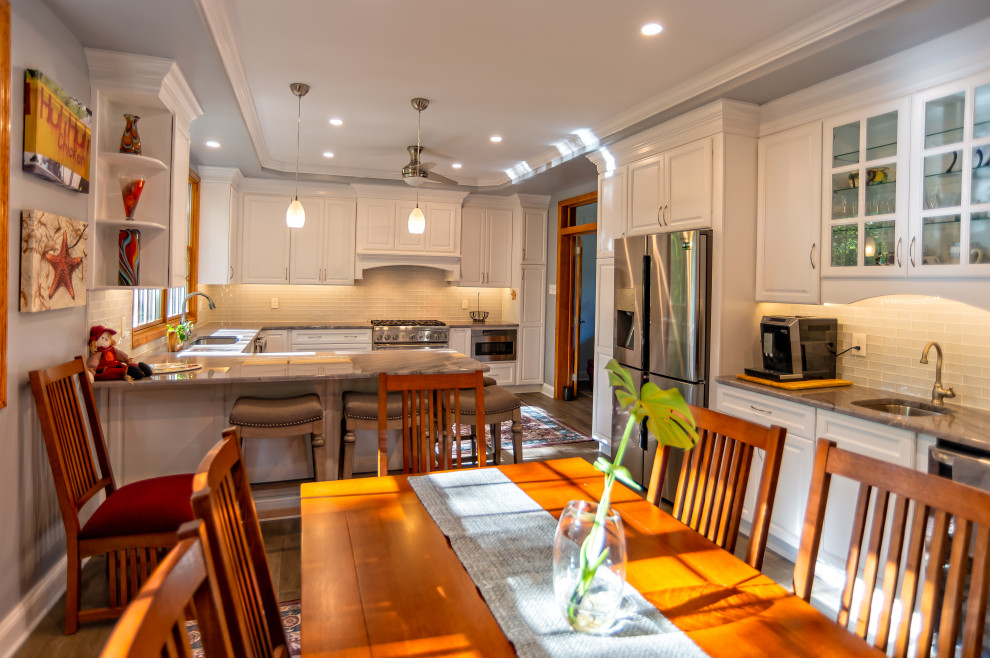Photo of a large traditional u-shaped eat-in kitchen in Philadelphia with an undermount sink, raised-panel cabinets, white cabinets, quartz benchtops, white splashback, glass tile splashback, stainless steel appliances, medium hardwood floors, a peninsula, brown floor and brown benchtop.