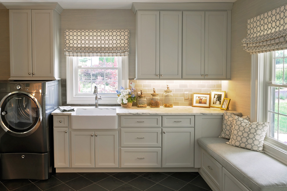 Inspiration for a large traditional single-wall utility room in New York with a farmhouse sink, recessed-panel cabinets, grey cabinets, granite benchtops, grey walls, porcelain floors and a side-by-side washer and dryer.