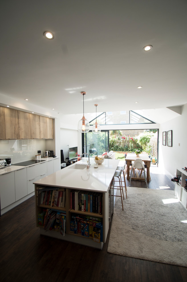 This is an example of a small modern open plan kitchen with an integrated sink, flat-panel cabinets, white cabinets, solid surface benchtops, white splashback, glass sheet splashback, stainless steel appliances, plywood floors, with island, brown floor and white benchtop.