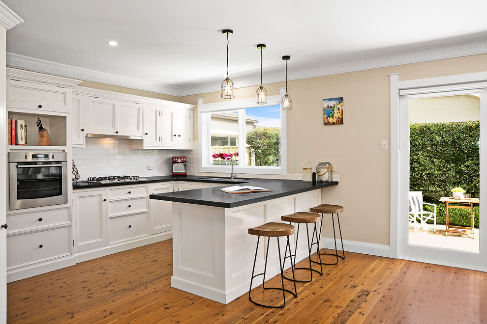 This is an example of a mid-sized transitional u-shaped eat-in kitchen in Sydney with a single-bowl sink, shaker cabinets, white cabinets, granite benchtops, white splashback, subway tile splashback, stainless steel appliances and medium hardwood floors.
