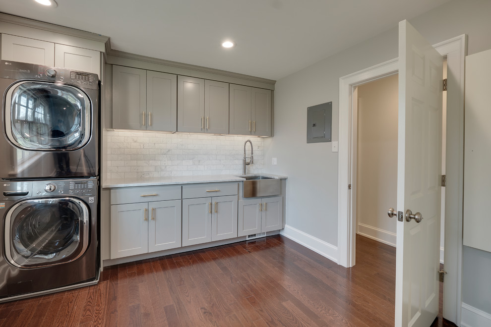 Mid-sized transitional single-wall dedicated laundry room in Philadelphia with a farmhouse sink, recessed-panel cabinets, grey cabinets, marble benchtops, grey walls, medium hardwood floors, a stacked washer and dryer, brown floor and white benchtop.