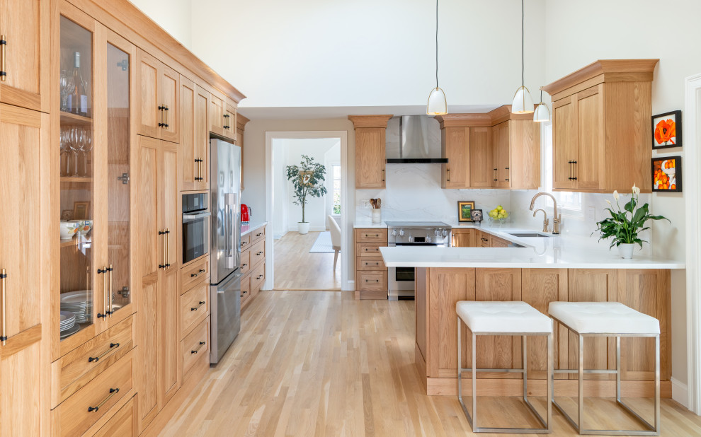 Inspiration for a transitional u-shaped light wood floor and beige floor kitchen remodel in Boston with an undermount sink, shaker cabinets, medium tone wood cabinets, white backsplash, stone slab backsplash, stainless steel appliances, a peninsula and white countertops