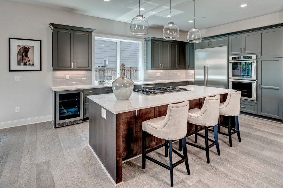 Large transitional l-shaped eat-in kitchen in Seattle with recessed-panel cabinets, grey cabinets, quartz benchtops, white splashback, ceramic splashback, stainless steel appliances, light hardwood floors, with island, grey floor and white benchtop.
