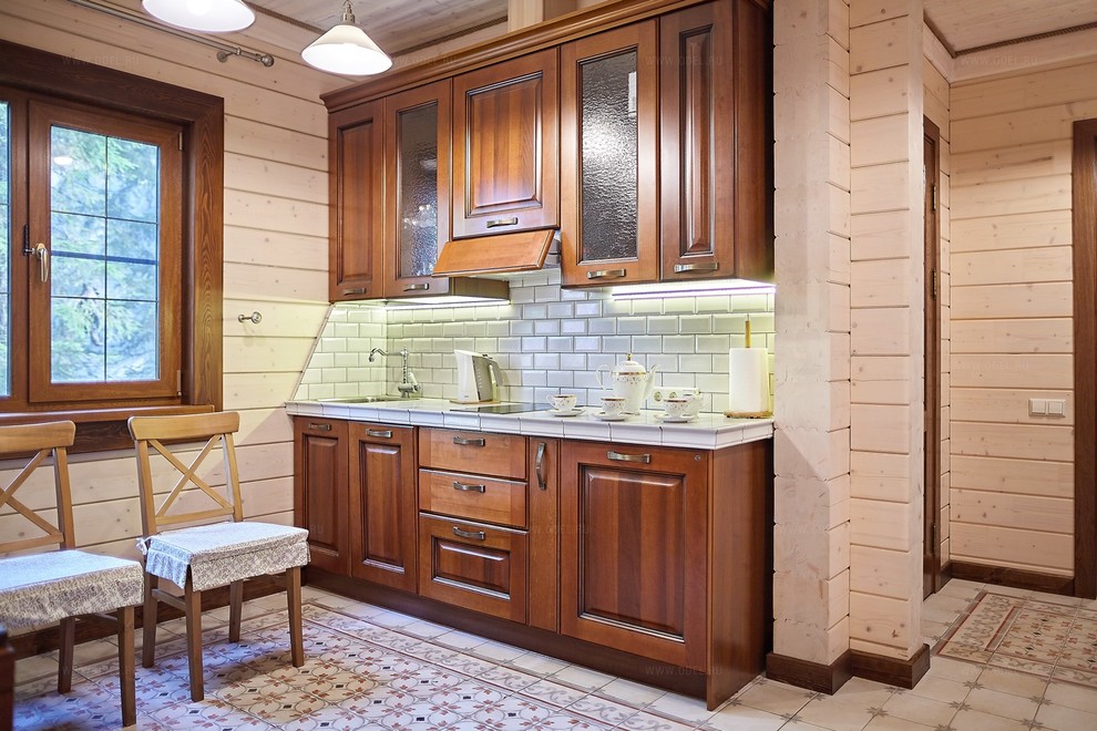 This is an example of a mid-sized country single-wall separate kitchen in Moscow with a drop-in sink, raised-panel cabinets, brown cabinets, tile benchtops, white splashback, ceramic splashback, panelled appliances, brick floors, no island, beige floor, white benchtop and wood.