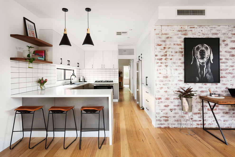 Photo of a mid-sized contemporary u-shaped kitchen in Melbourne with flat-panel cabinets, white cabinets, white splashback, ceramic splashback, medium hardwood floors, brown floor, white benchtop, a drop-in sink, panelled appliances and a peninsula.