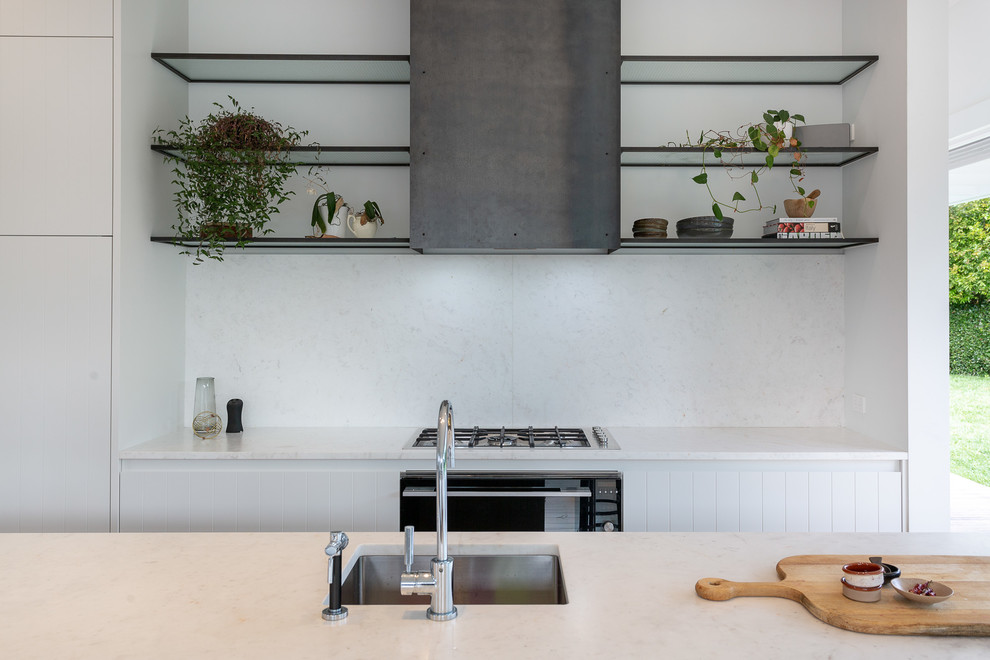 Mid-sized modern galley open plan kitchen in Auckland with a drop-in sink, white cabinets, marble benchtops, white splashback, marble splashback, black appliances, light hardwood floors, with island, brown floor and white benchtop.