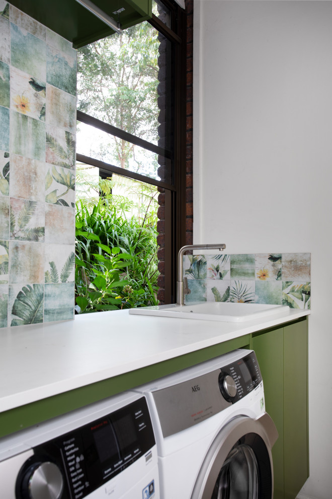 Kleiner Moderner Hauswirtschaftsraum in L-Form mit Einbauwaschbecken, grünen Schränken, Quarzwerkstein-Arbeitsplatte, Küchenrückwand in Grün, Rückwand aus Porzellanfliesen, bunten Wänden, Porzellan-Bodenfliesen, Waschmaschine und Trockner nebeneinander, grauem Boden und weißer Arbeitsplatte in Brisbane