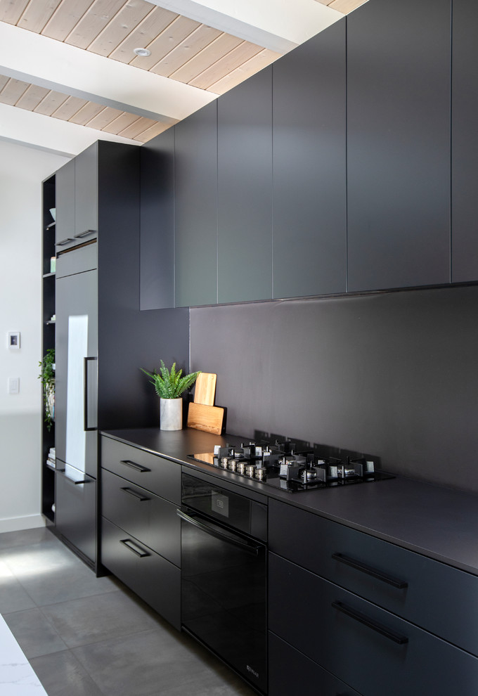 Photo of a large midcentury single-wall open plan kitchen in Vancouver with flat-panel cabinets, black cabinets, quartz benchtops, black splashback, with island, grey floor, black benchtop, an undermount sink, panelled appliances and concrete floors.