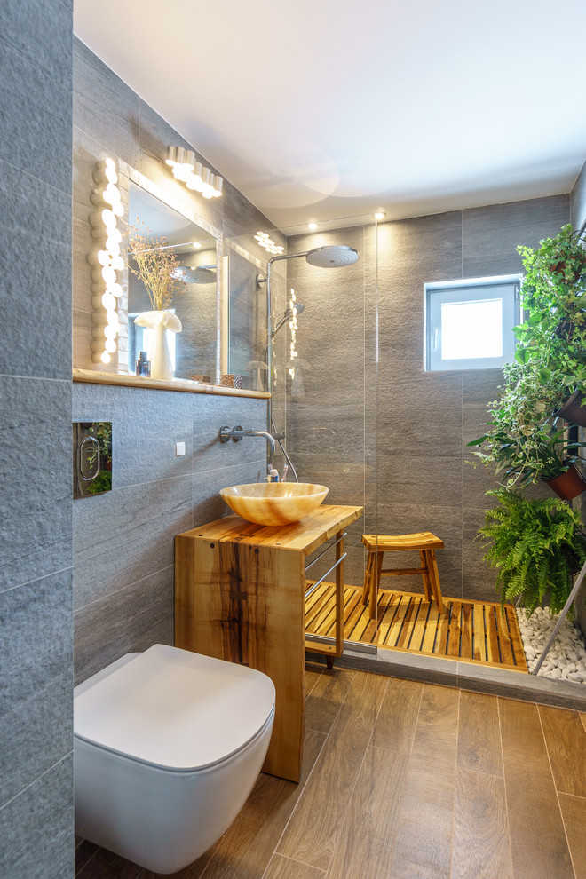 This is an example of a small tropical 3/4 bathroom in Other with white cabinets, an alcove shower, a wall-mount toilet, gray tile, ceramic tile, grey walls, ceramic floors, a vessel sink and wood benchtops.
