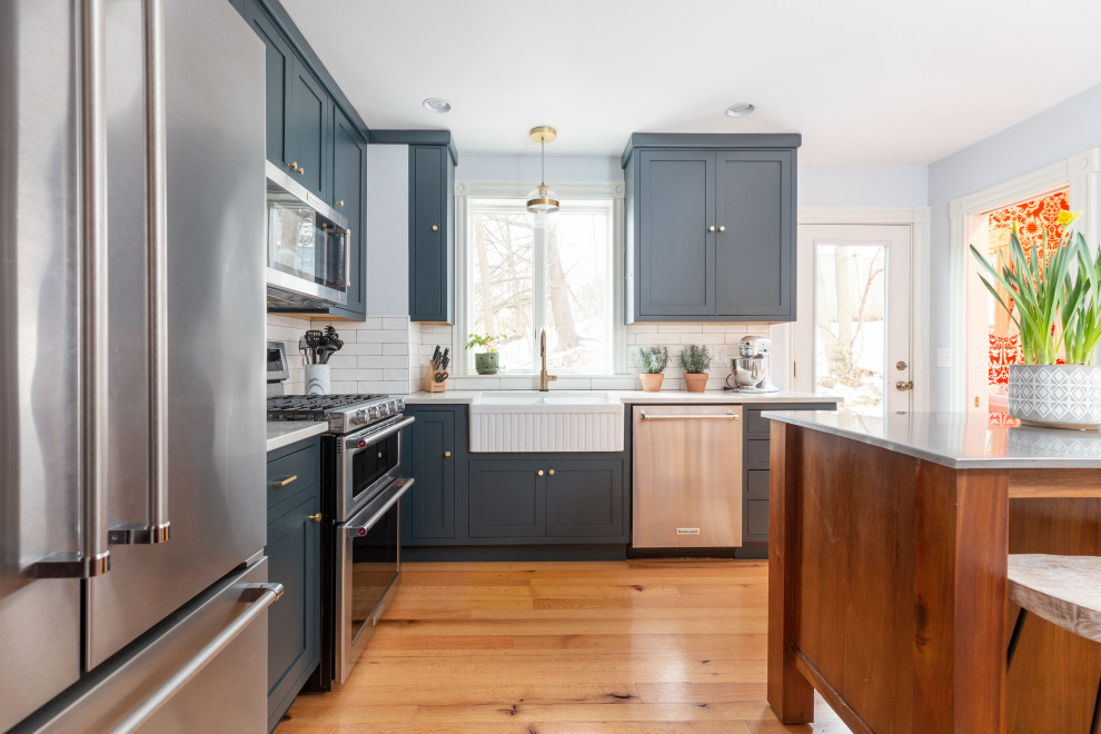 Inspiration for a small transitional l-shaped separate kitchen in Cleveland with a farmhouse sink, shaker cabinets, blue cabinets, quartz benchtops, white splashback, ceramic splashback, stainless steel appliances, light hardwood floors, with island and white benchtop.