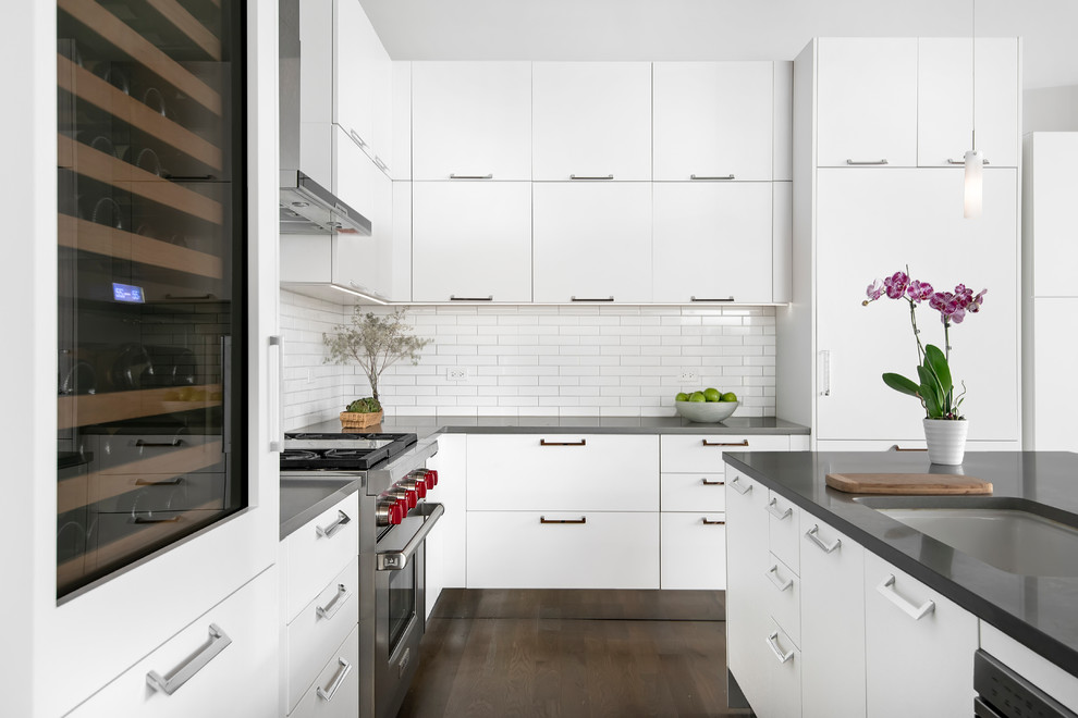 This is an example of an industrial kitchen in Chicago with white cabinets, white splashback, stainless steel appliances, with island, brown floor and grey benchtop.