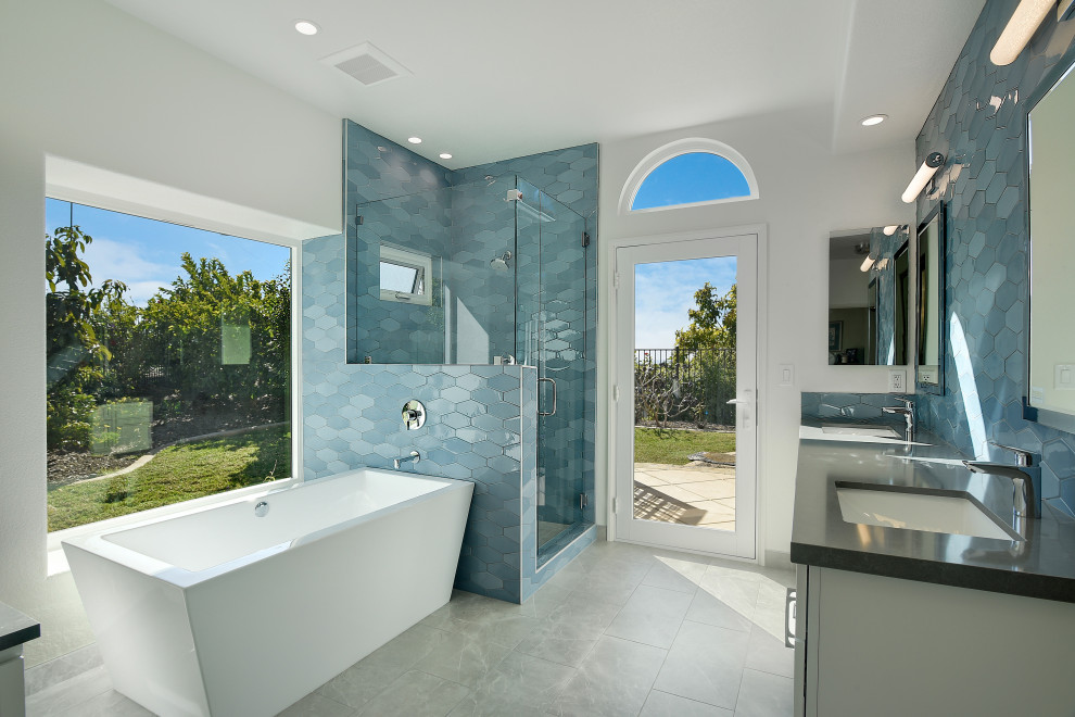 Photo of a mid-sized contemporary master bathroom in San Diego with shaker cabinets, white cabinets, a freestanding tub, a corner shower, a one-piece toilet, blue tile, ceramic tile, white walls, ceramic floors, an undermount sink, engineered quartz benchtops, grey floor, a hinged shower door, grey benchtops, a niche, a double vanity and a built-in vanity.