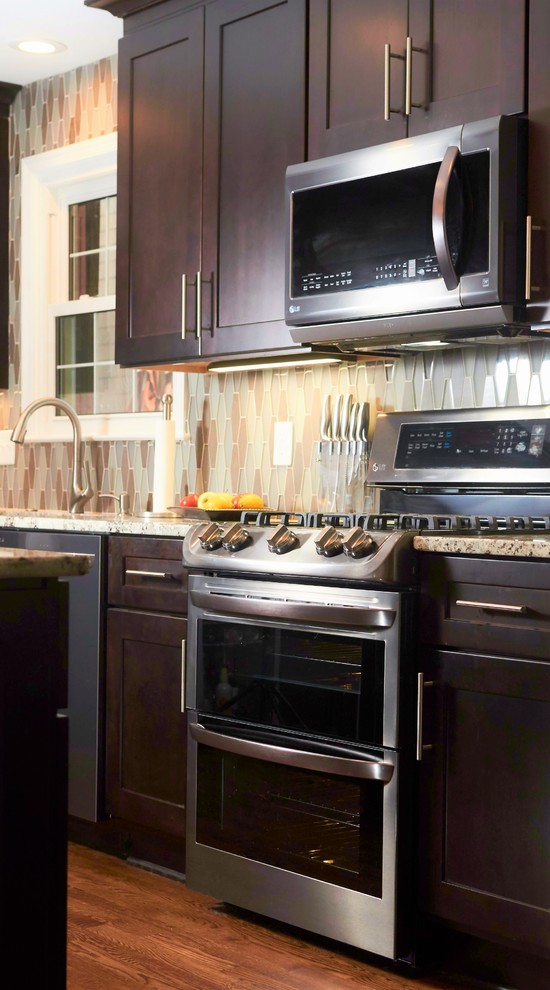 Transitional l-shaped medium tone wood floor and brown floor open concept kitchen photo with an undermount sink, shaker cabinets, dark wood cabinets, granite countertops, multicolored backsplash, mosaic tile backsplash, stainless steel appliances, an island and white countertops