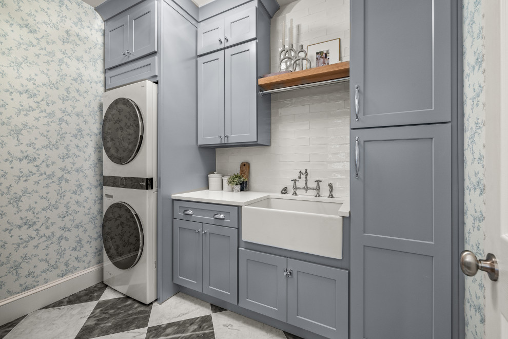 Inspiration for a medium sized traditional separated utility room in Atlanta with a belfast sink, shaker cabinets, grey cabinets, engineered stone countertops, white splashback, metro tiled splashback, blue walls, marble flooring, a stacked washer and dryer, multi-coloured floors, white worktops and wallpapered walls.