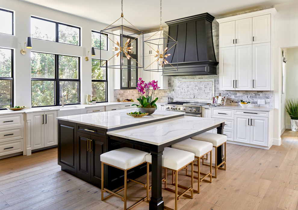 Photo of a large traditional u-shaped kitchen in Austin with white cabinets, quartz benchtops, marble splashback, stainless steel appliances, light hardwood floors, with island, white benchtop and raised-panel cabinets.
