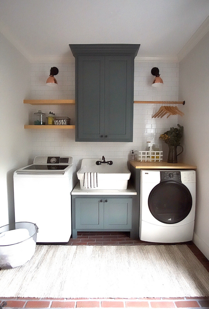This is an example of a mid-sized country galley dedicated laundry room in Philadelphia with a farmhouse sink, shaker cabinets, green cabinets, quartz benchtops, grey walls, brick floors, a side-by-side washer and dryer, multi-coloured floor and white benchtop.