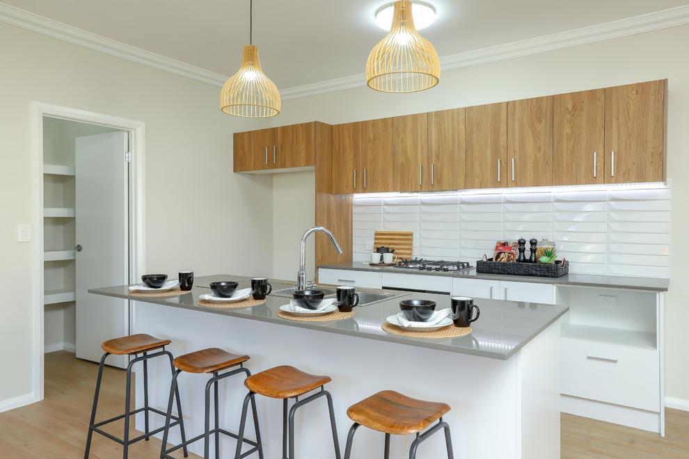 Photo of a mid-sized transitional galley open plan kitchen in Newcastle - Maitland with a double-bowl sink, medium wood cabinets, quartz benchtops, white splashback, ceramic splashback, stainless steel appliances, vinyl floors, with island, brown floor and grey benchtop.
