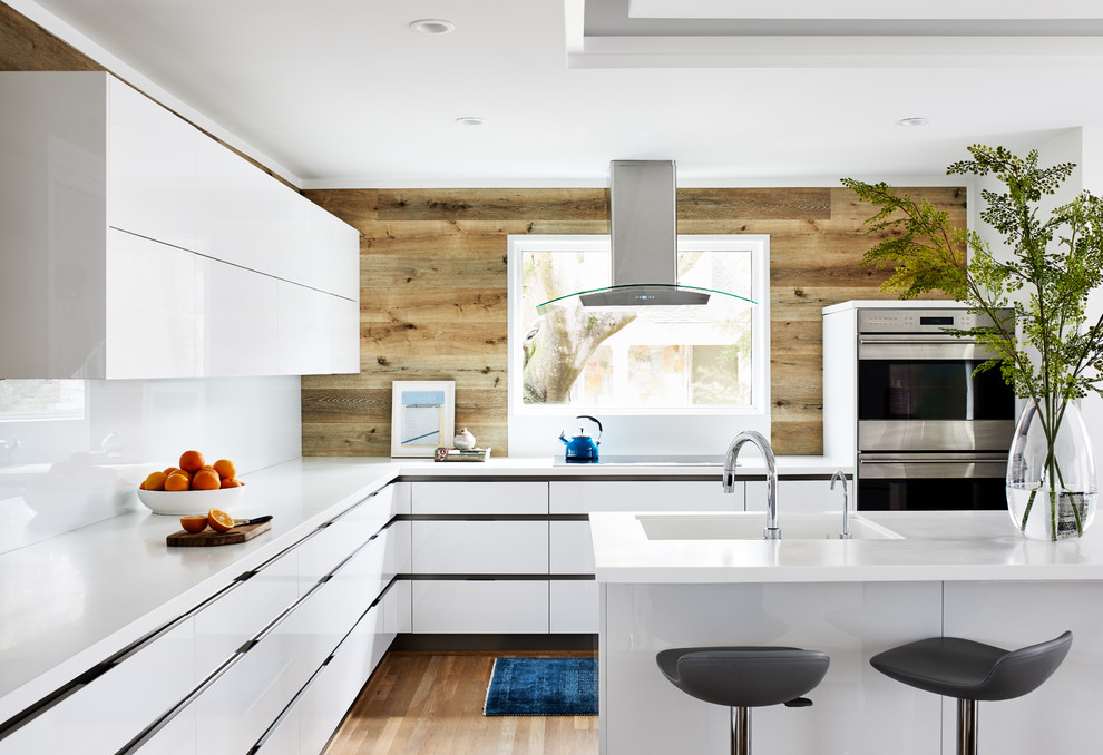 Inspiration for a mid-sized midcentury u-shaped kitchen in DC Metro with flat-panel cabinets, white cabinets, solid surface benchtops, white splashback, stainless steel appliances, with island, white benchtop, an integrated sink, medium hardwood floors and brown floor.