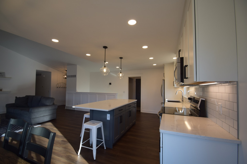 Example of a mid-sized transitional single-wall vinyl floor open concept kitchen design with a farmhouse sink, shaker cabinets, quartz countertops, white backsplash, subway tile backsplash, stainless steel appliances, an island and white countertops