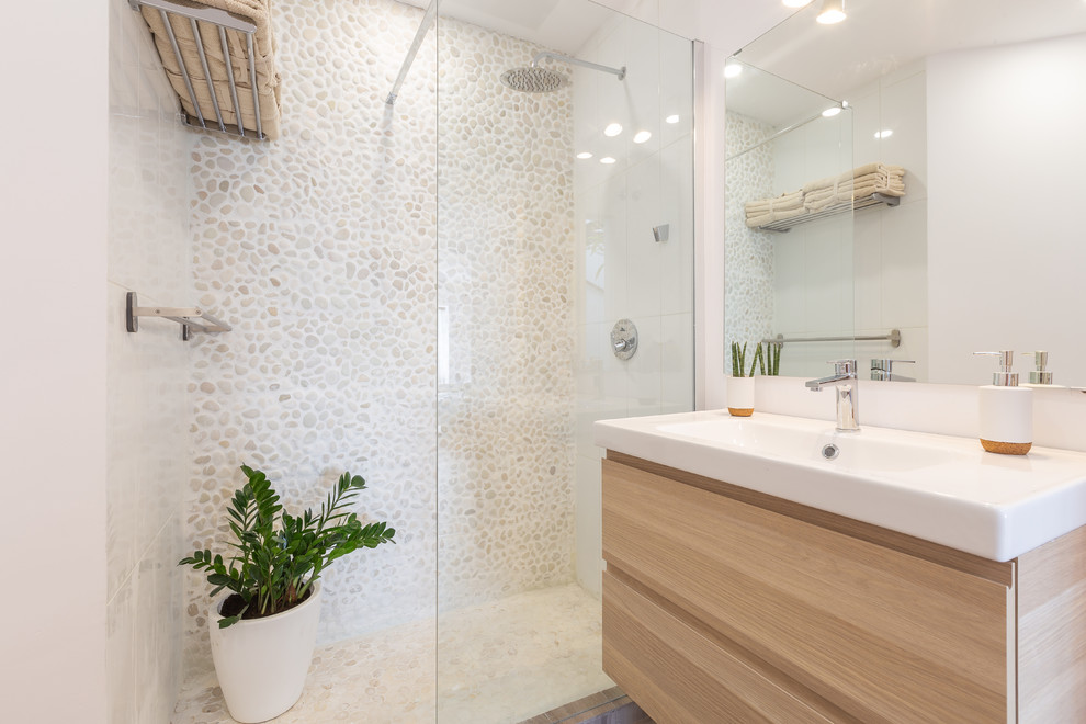 This is an example of a mid-sized mediterranean 3/4 bathroom in Malaga with a curbless shower, white walls, white benchtops, flat-panel cabinets, light wood cabinets, beige tile, pebble tile, an integrated sink and an open shower.